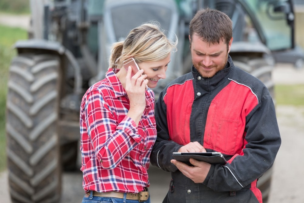 Man and woman consulting a digital tablet, woman making a phone call from it. A tractor can be seen in the background. Man and woman consulting a digital tablet, woman making a phone call from it. A tractor can be seen in the background.